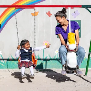 An All Hands and Hearts staff member and young student play on a swing set, smiling at one another