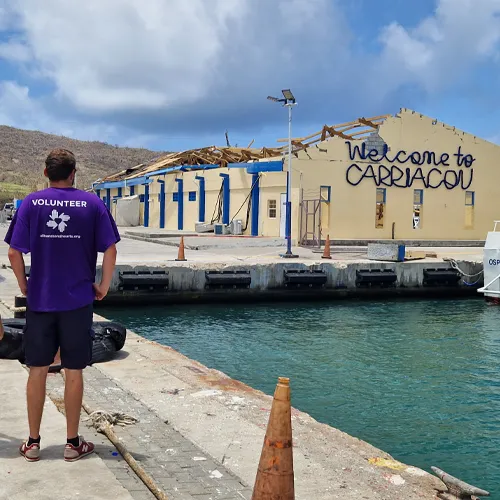 Hurricane Beryl relief volunteer stands at Carriacou dock in front of a storm-damaged building in Grenada.
