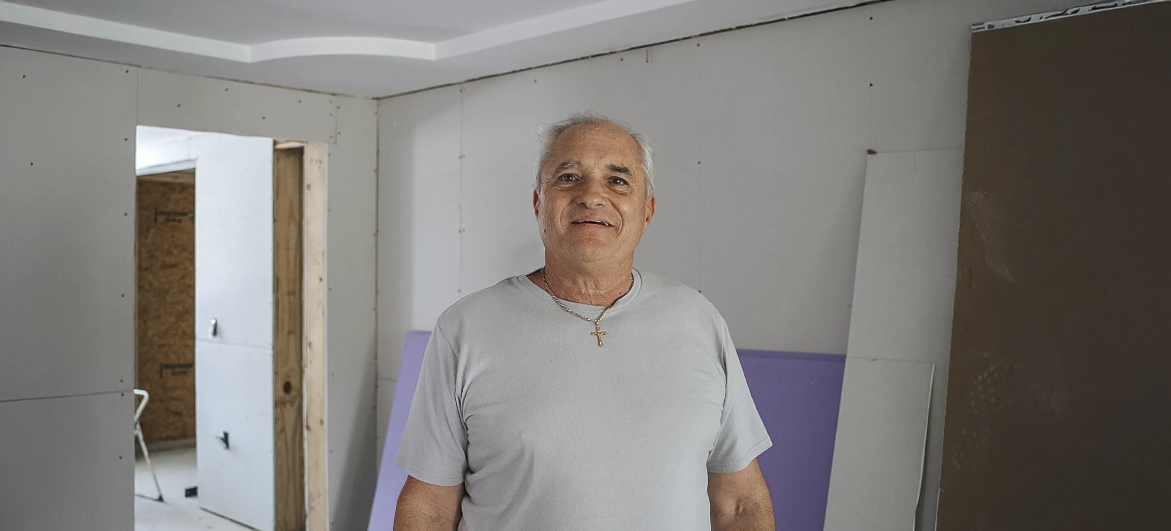 Un hombre se encuentra en el interior de una casa parcialmente reconstruida, sonriendo levemente, con paredes inacabadas y materiales de construcción visibles a su alrededor.