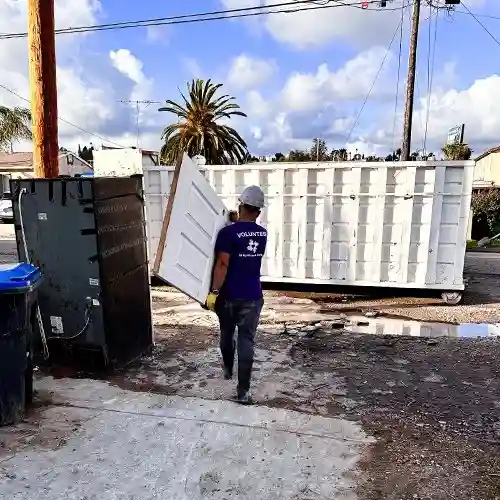 Volunteer in a purple All Hands and Hearts shirt carries a damaged door toward a large debris container during California flood cleanup.
