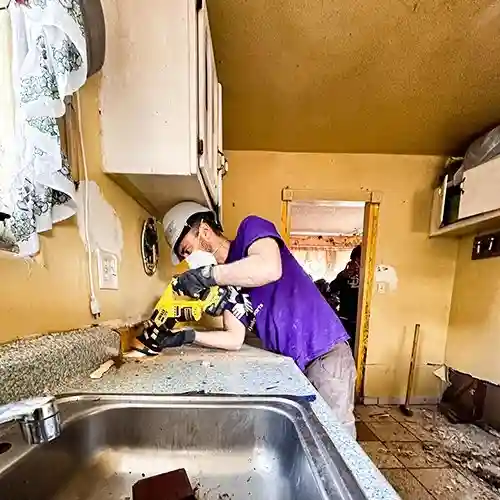 Volunteer in a hard hat and safety glasses clears debris inside a kitchen damaged by California flooding.