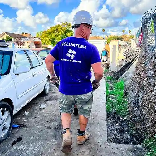 All Hands and Hearts volunteer in a hard hat and safety gear carries tools through a flood-affected neighborhood during cleanup efforts.