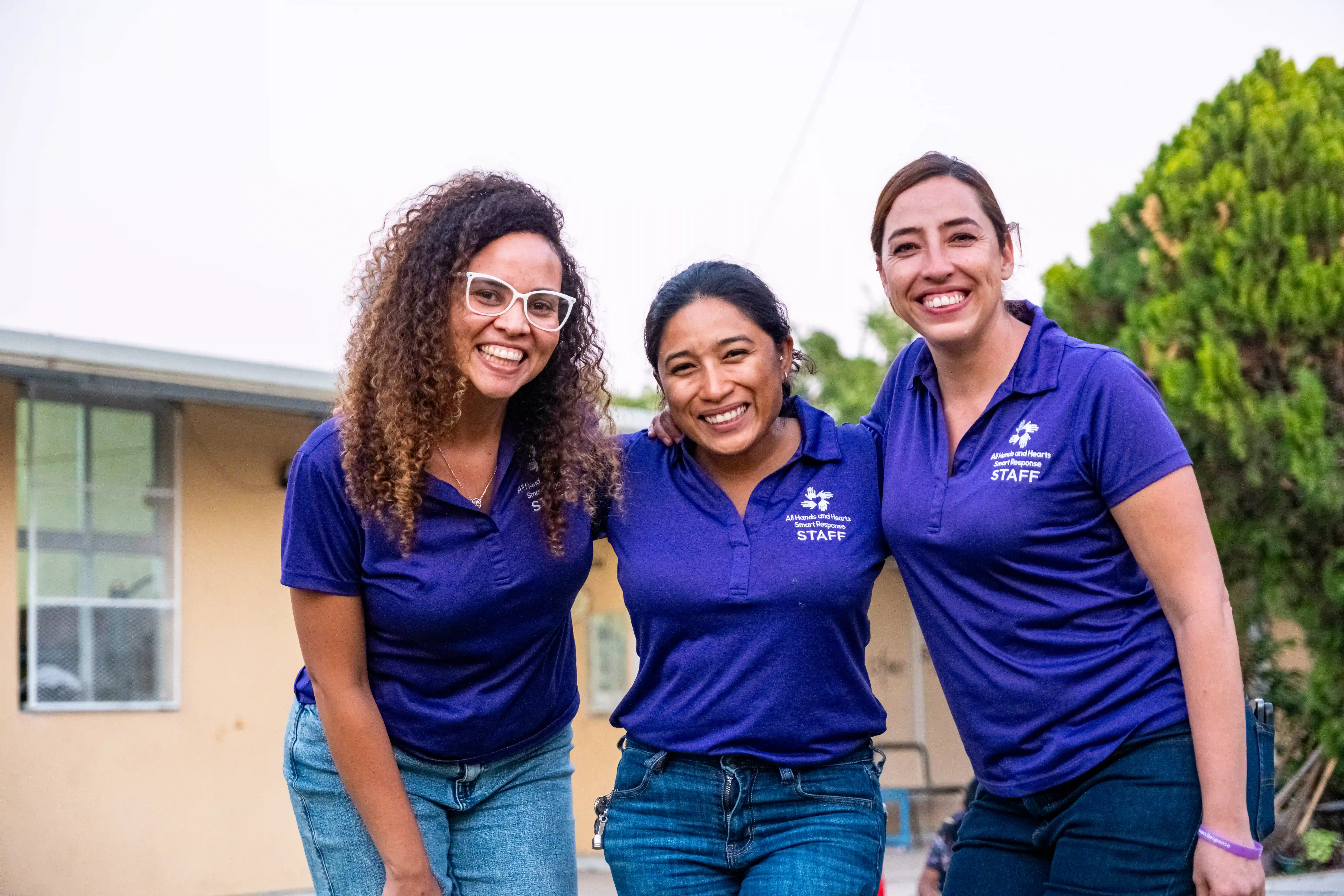 Tres mujeres vestidas con polos morados a juego con el logotipo de All Hands &amp; Hearts permanecen juntas al aire libre, sonriendo cariñosamente a la cámara.
