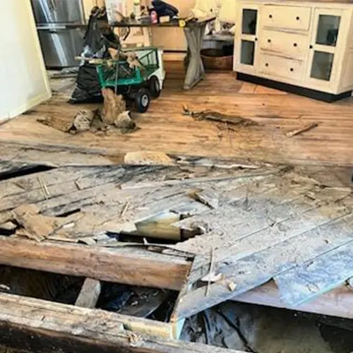 The wooden floor inside a house is torn apart with visible debris and water damage.