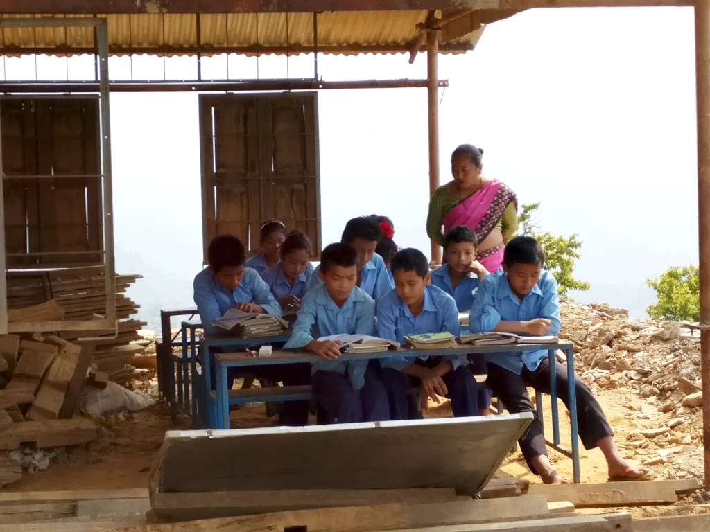 A group of children in blue school uniforms sit at desks studying outdoors under a makeshift shelter with wooden beams and a tin roof, while a teacher stands beside them.