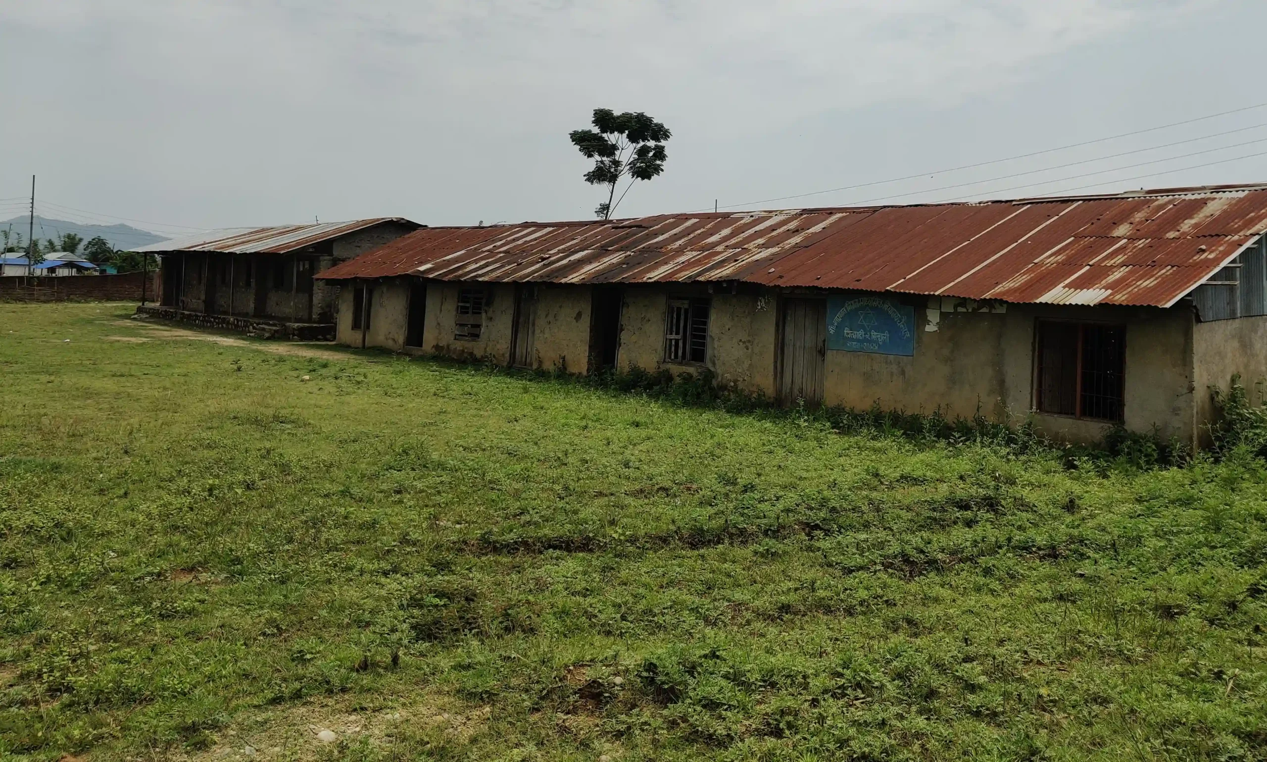 An old, worn-down school building with a rusted metal roof and faded walls, standing beside an open grassy field.