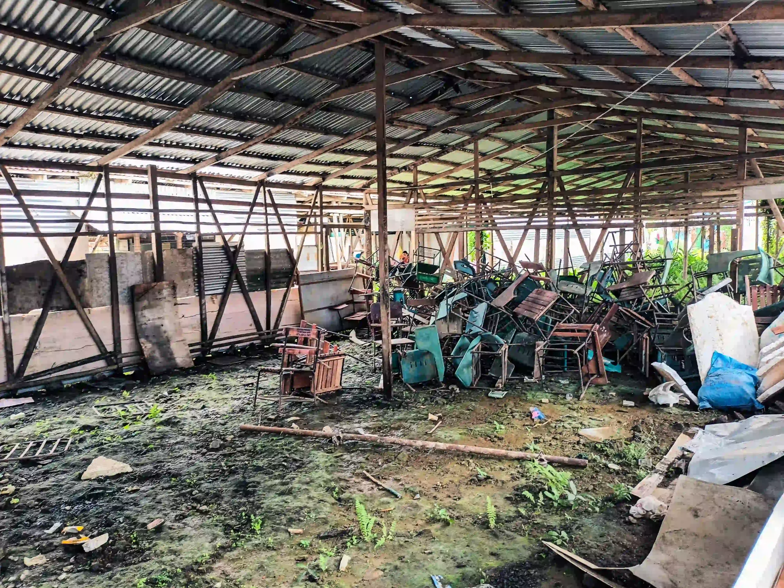 A damaged school building with a rusted metal roof, broken wooden frame, and piles of overturned desks and debris on a muddy floor.