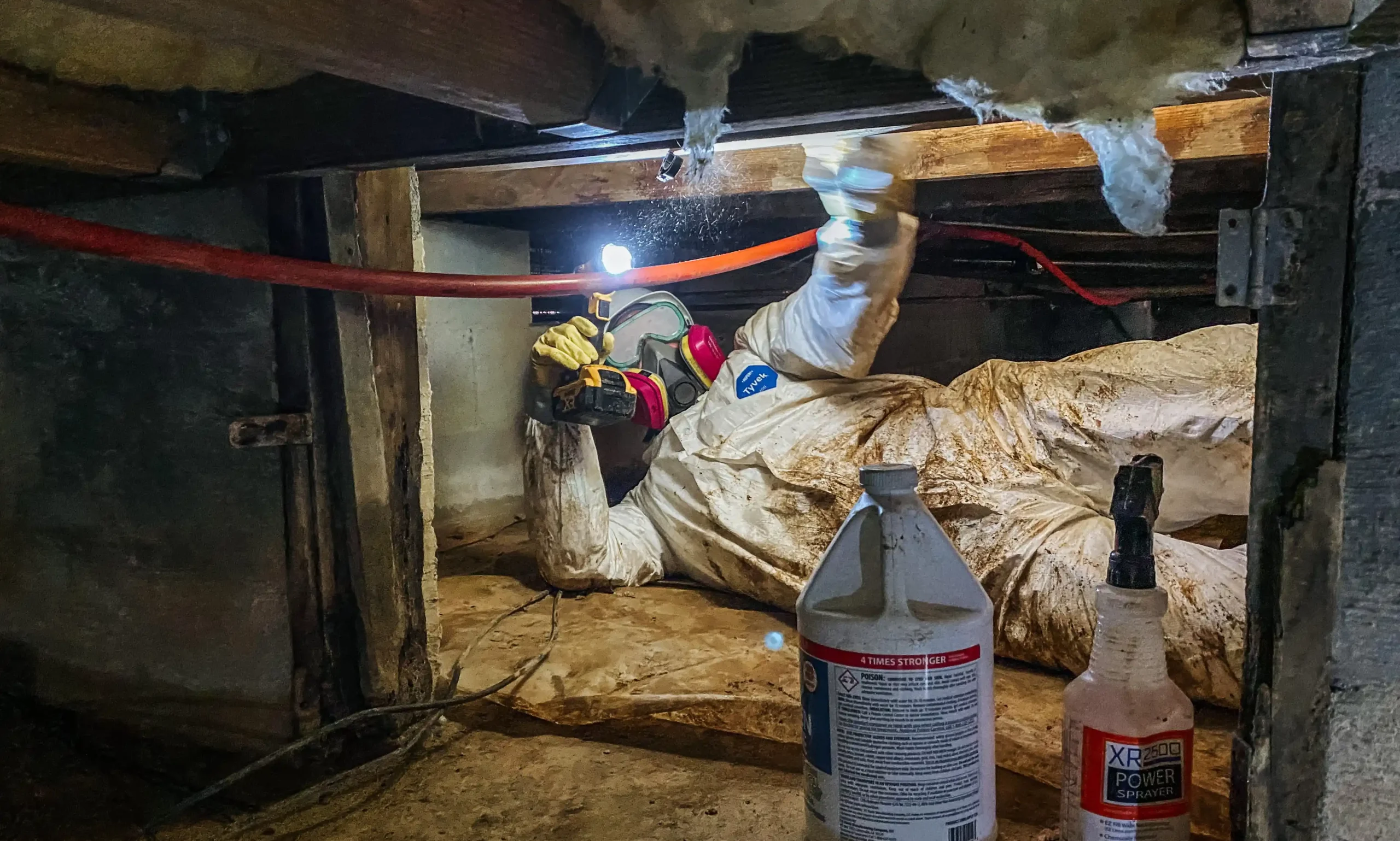 An All Hands and Hearts volunteer in a tyvek suit, hard hat, goggles and respirator in a small crawl space of a home spraying for mold sanitation