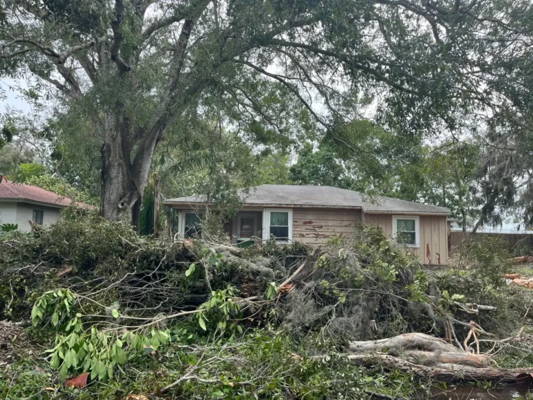 A damaged home with piles of tree debris surrounding the front of the home
