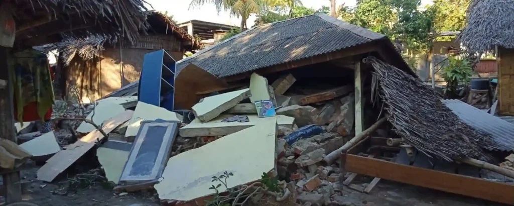 A collapsed house in Lombok with its roof caved in and walls reduced to rubble, debris and broken furniture scattered across the ground.