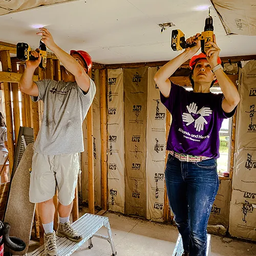 Two volunteers in safety gear use drills to attach drywall to a ceiling inside a storm-damaged house.