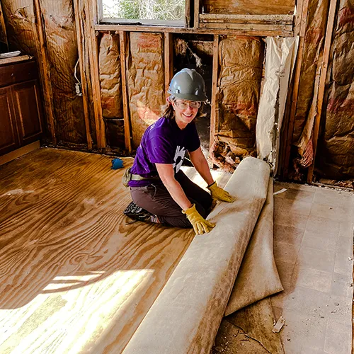 A smiling volunteer in a hard hat and gloves kneels inside a damaged home, pulling up old flooring.