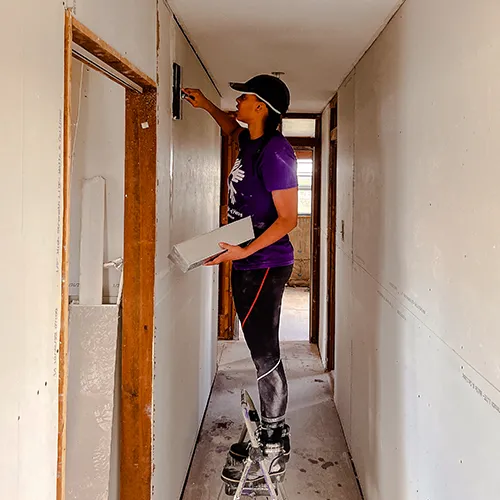 A volunteer stands on a step stool applying joint compound to drywall seams in a hallway of a rebuilt home.