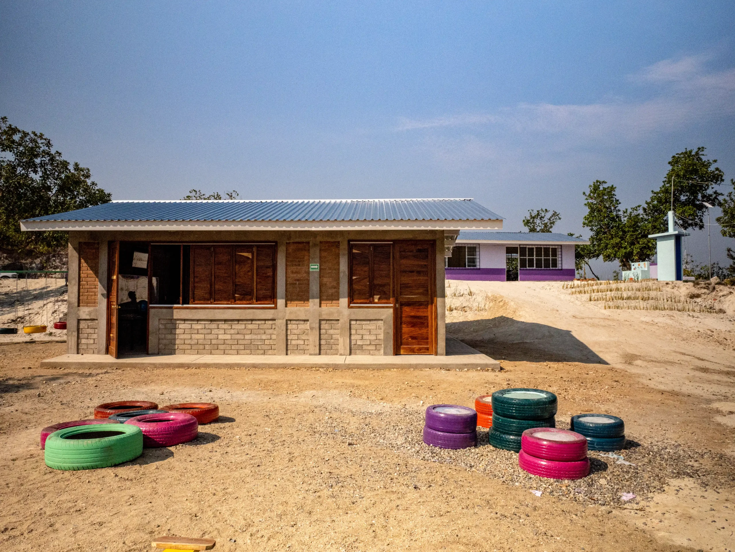 A newly rebuilt school with sturdy brick walls, a metal roof, and a play area with colorful tire seats in front.