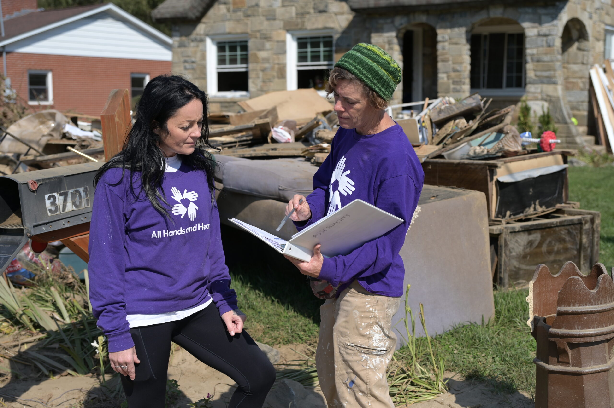Dos voluntarios de All Hands &amp; Hearts, de pie frente a enormes montones de escombros domésticos con un portapapeles, realizan evaluaciones de viviendas.