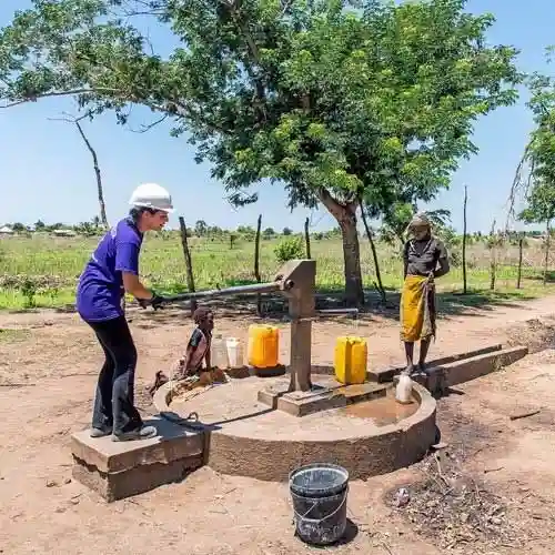 An All Hands and Hearts volunteer operates a hand water pump while a woman waits with yellow jerry cans.