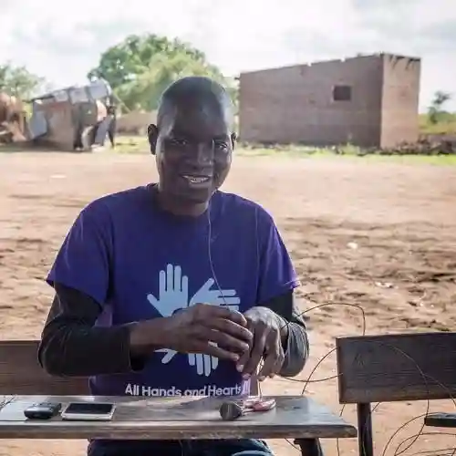 A local man wearing an All Hands and Hearts shirt smiles while seated at a wooden desk outdoors.