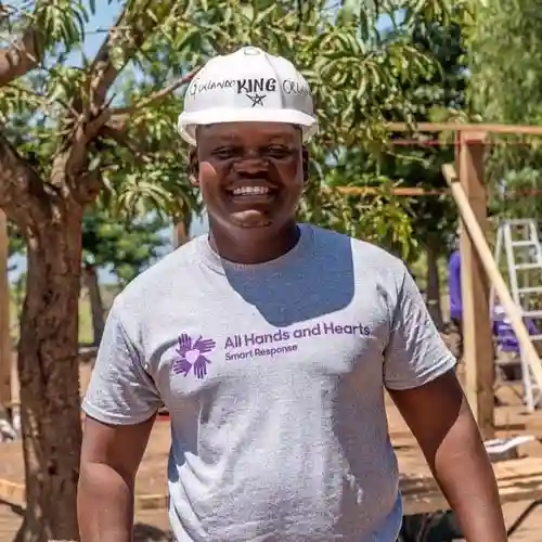 A smiling man in a white hard hat and All Hands and Hearts shirt stands outdoors at a construction site.