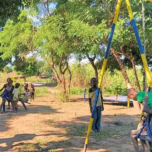 Children play on swings and a seesaw in an outdoor area surrounded by trees in Mozambique.