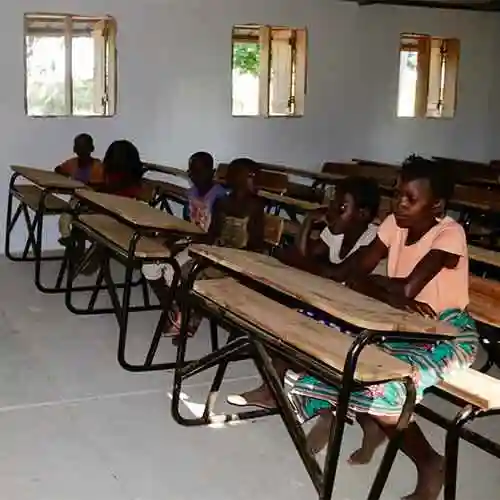Students sit at wooden desks inside a simple classroom with white walls and open windows.