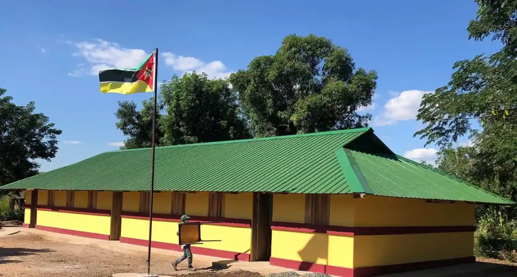 A child walks past a newly built school painted yellow and red with a green roof and Mozambique flag.