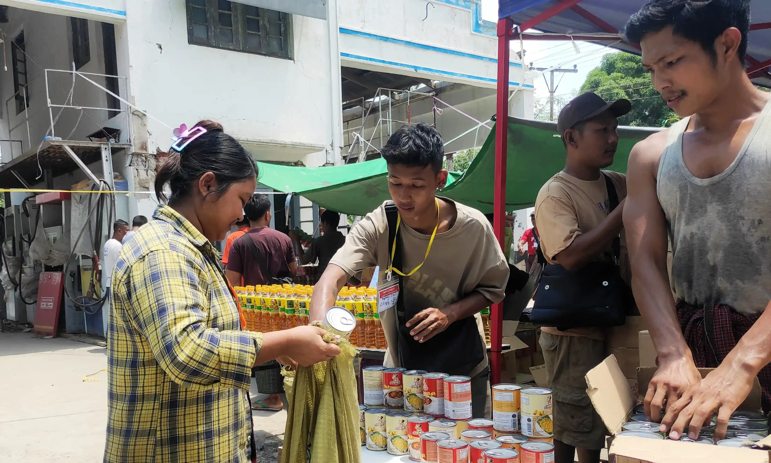Community members in Myanmar distribute canned food and supplies at an outdoor relief station, with people lining up to receive aid.
