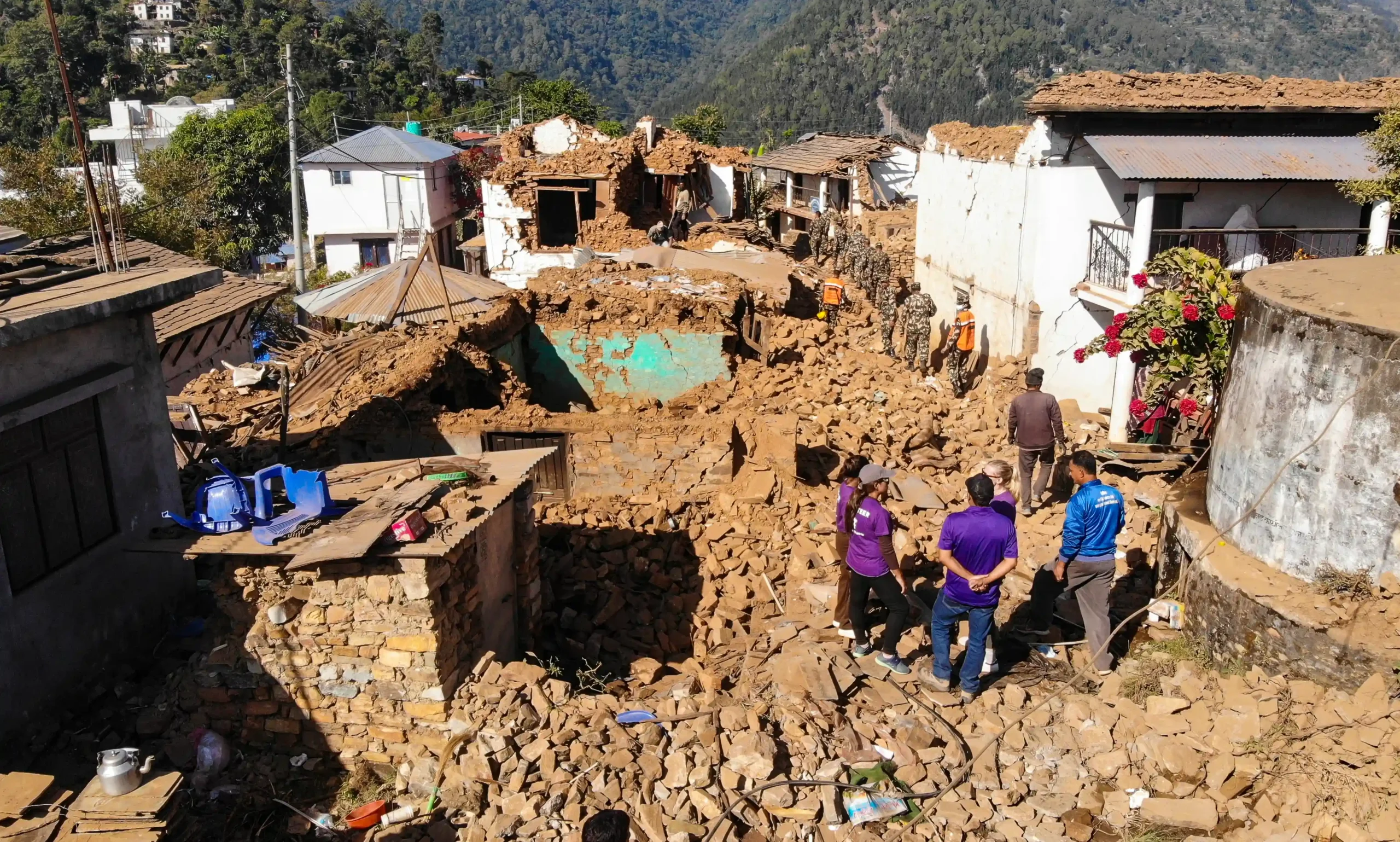 An earthquake-damaged neighborhood in Nepal, with collapsed homes reduced to rubble. A small group of people, including volunteers in purple shirts, stand among the destruction.