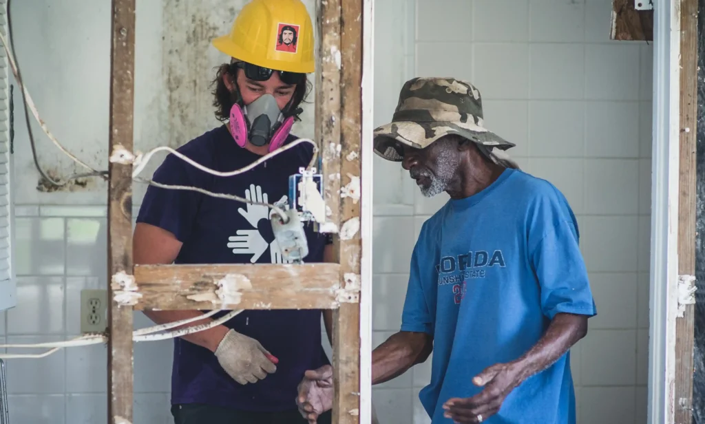 A volunteer works beside a community member in a blue shirt and hat inside a damaged home, with exposed wall framing and wires visible.