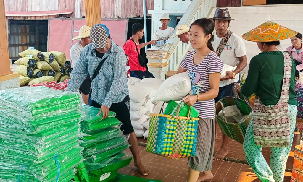 Community members carry baskets and bags of supplies while unloading and organizing relief goods, including stacked green packages, in a distribution area.