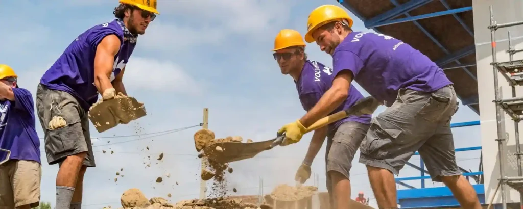 os voluntarios con camisetas moradas de All Hands and Hearts y cascos amarillos palean tierra juntos en una obra en Perú, trabajando en la reconstrucción tras el terremoto.