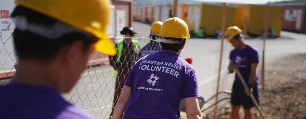 Un grupo de voluntarios con camisetas moradas y cascos amarillos instalan vallas cerca de las viviendas provisionales en un centro asistencia post-desastre .