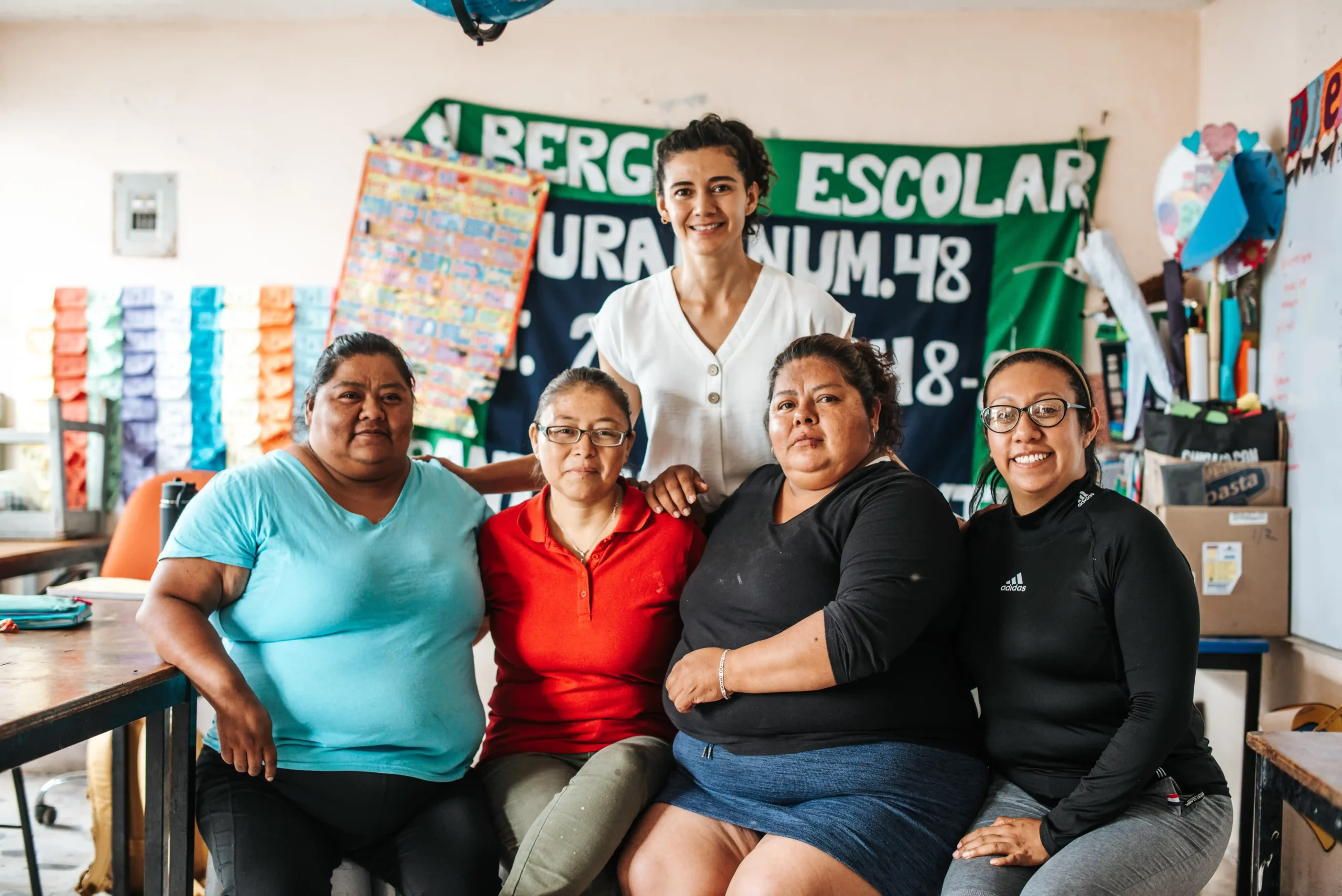 Un grupo de cinco mujeres se sientan juntas y sonríen a la cámara dentro de un aula, con una pancarta verde en la que se lee "Albergue Escolar" visible en la pared detrás de ellas.