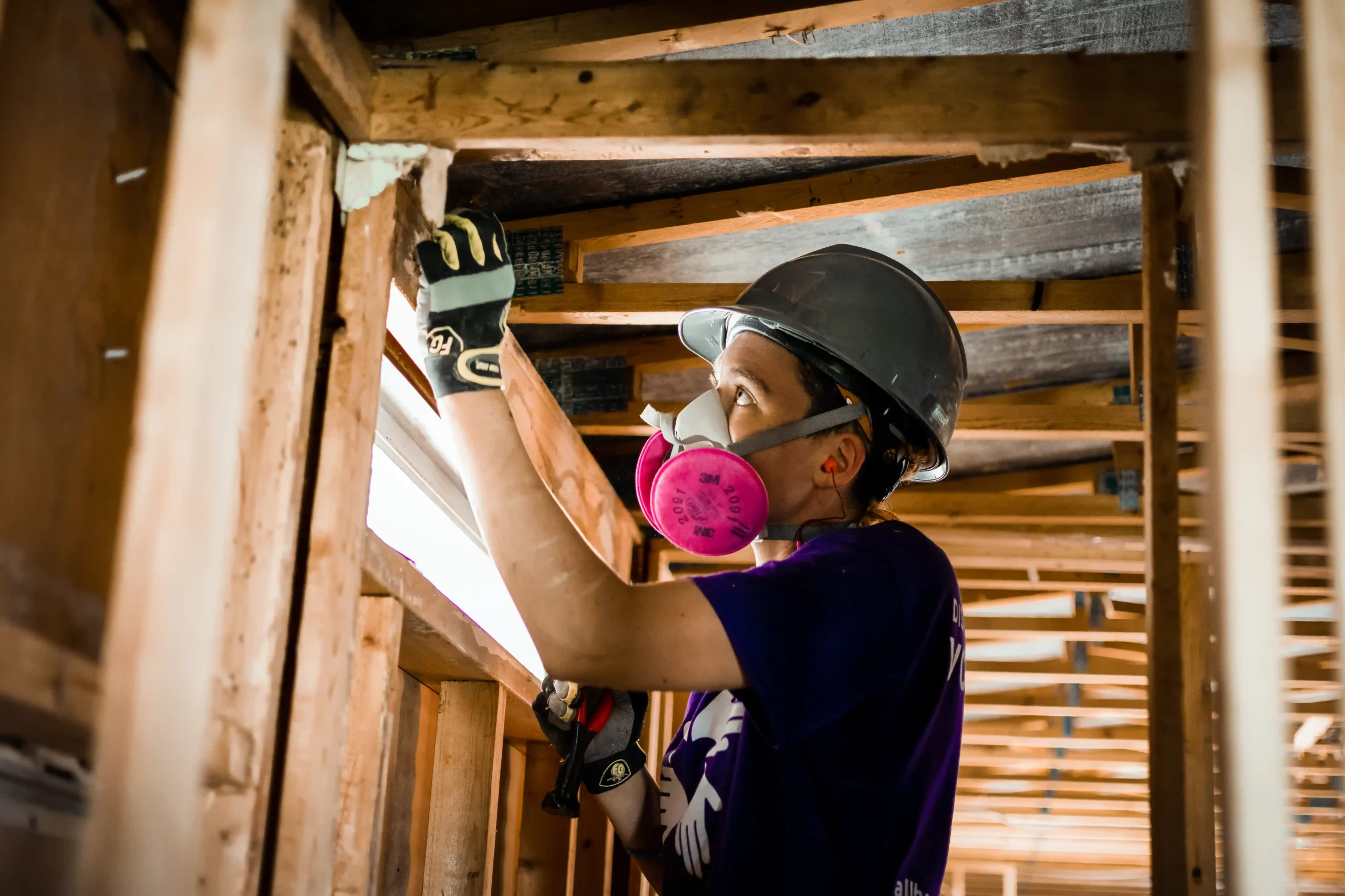 Un voluntario con casco, guantes y máscara protectora trabaja en el interior de la estructura de madera de una casa, utilizando una herramienta para reparar o fijar vigas.