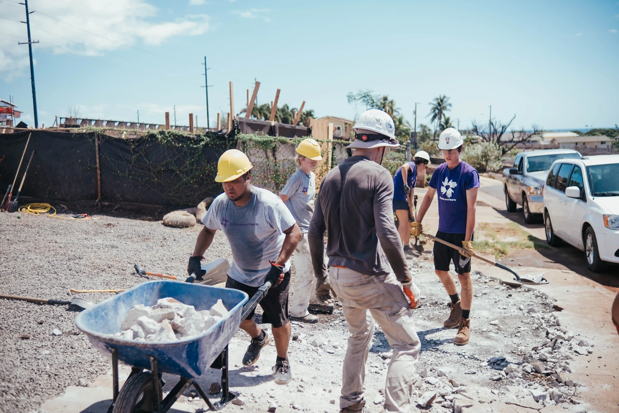 Voluntarios con casco y equipo de seguridad trabajan juntos al aire libre, retirando escombros y moviendo piedras con una carretilla en una zona afectada por una catástrofe.