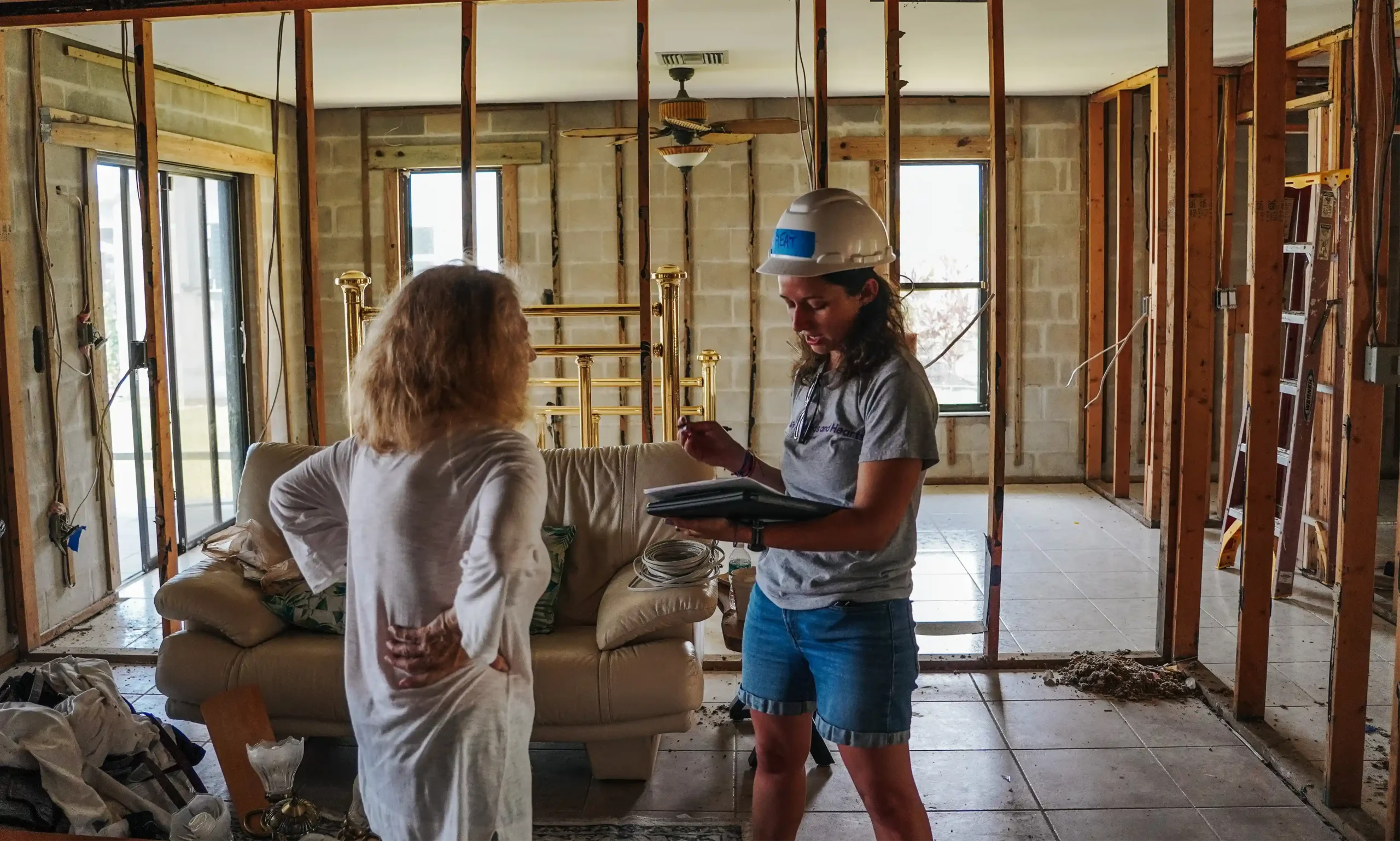 Un voluntario con casco y portapapeles habla con un propietario de una casa parcialmente dañada, en la que se ven paredes y materiales de construcción expuestos.