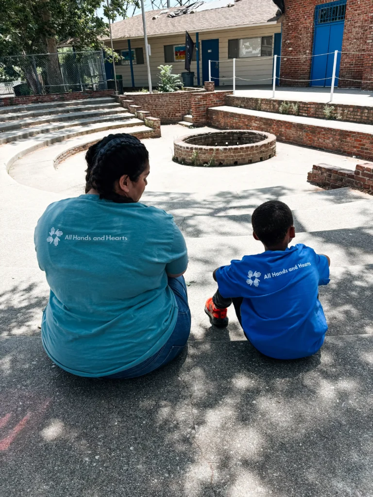 Una mujer y un niño, ambos con camisetas de All Hands and Hearts , sentados uno al lado del otro en el suelo del patio de un colegio, de espaldas a la cámara.