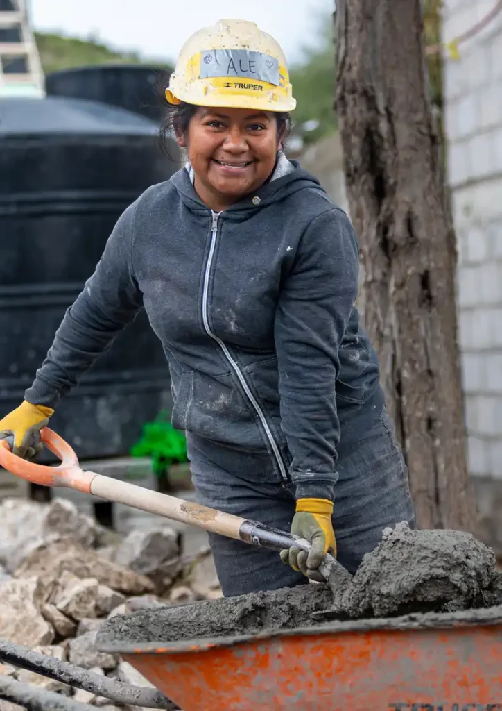 Alejandra sonríe a la cámara con una pala en la mano, moviendo cemento en las obras de reconstrucción de una escuela en México.