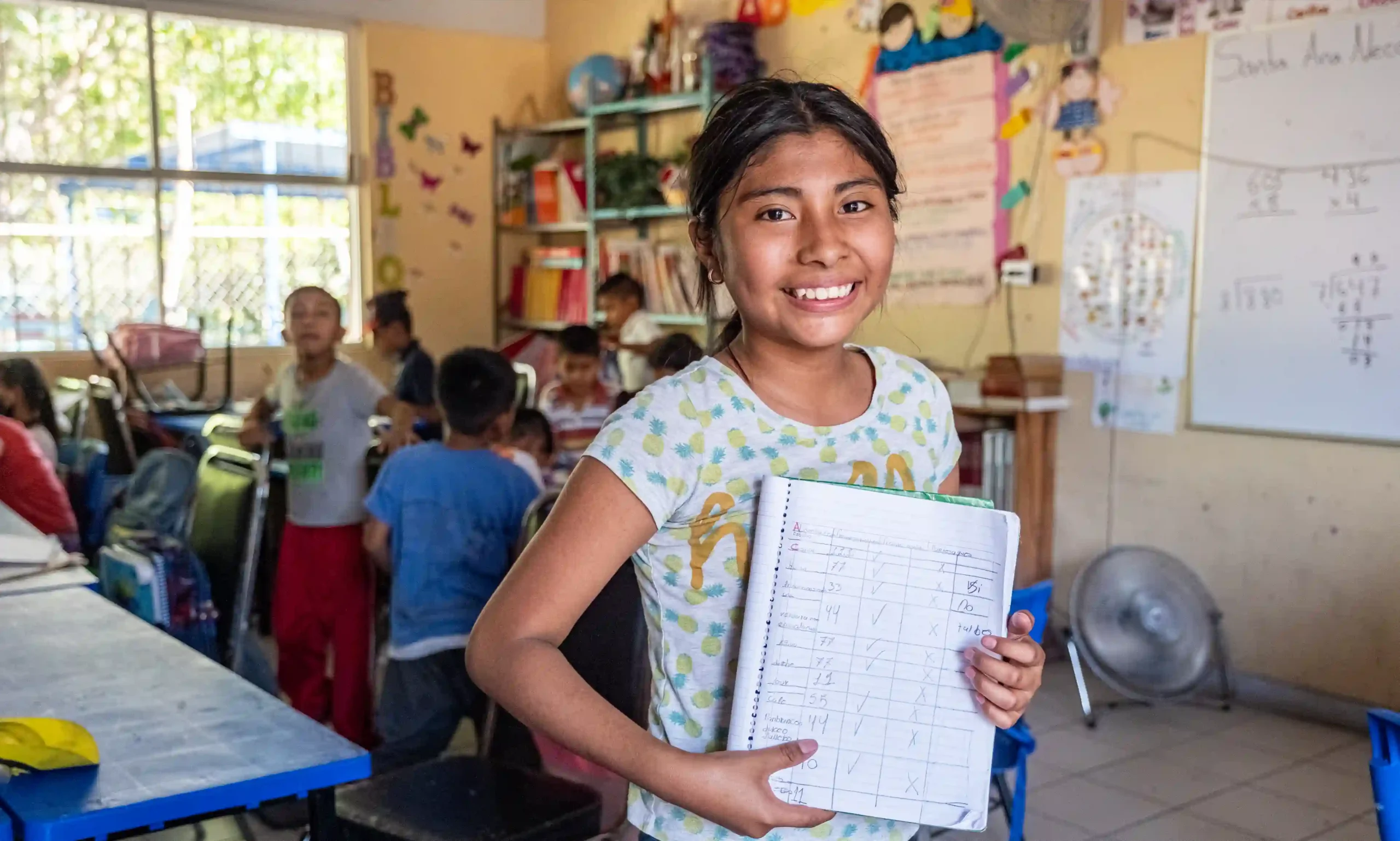 A smiling girl stands in a brightly decorated classroom holding up her notebook, while other children sit at desks behind her.
