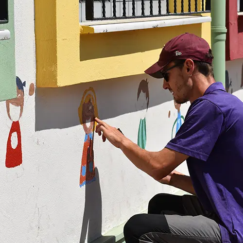 Un voluntario con gorra granate pinta alegres dibujos animados de niños en la pared de un edificio escolar.
