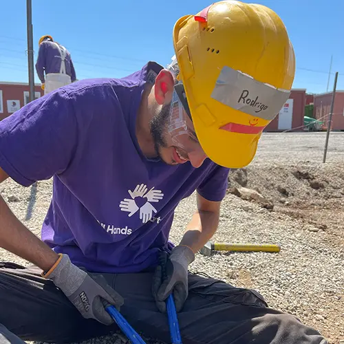 Un voluntario con equipo de seguridad y un casco amarillo con la etiqueta "Rodrigo" corta material de construcción con unos alicates en una parcela de grava.