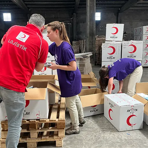 Voluntarios con camisetas moradas y un compañero con camiseta roja de logística organizan cajas de socorro etiquetadas con el logotipo de la Media Luna Roja Turca.