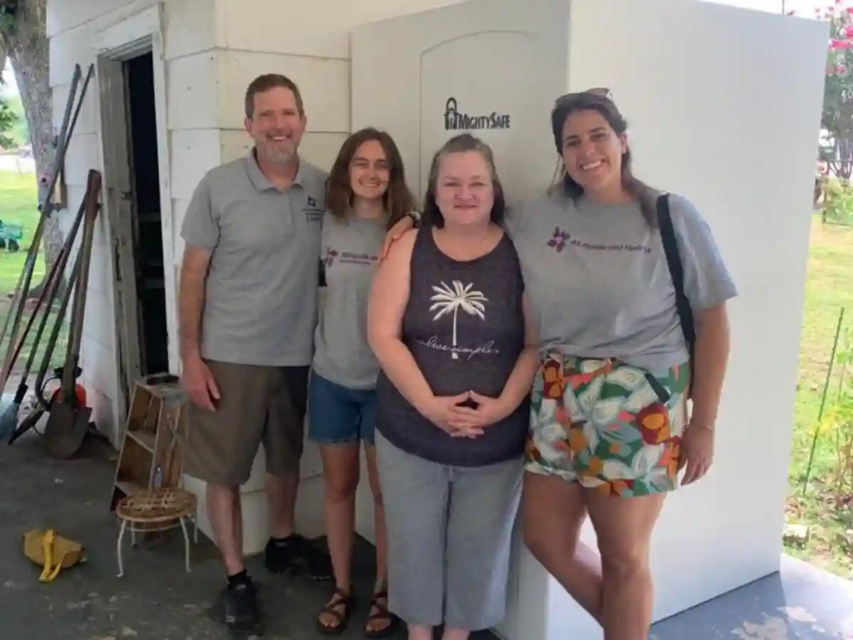 Volunteers and a homeowner stand smiling in front of a newly installed white storm shelter.