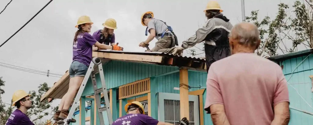 Team of volunteers in yellow hard hats repairing the roof of a turquoise wooden house in Puerto Rico as a local community member looks on.
