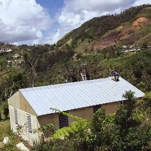 Volunteer repairing the roof of a hurricane-damaged home in Puerto Rico, with green hills and mountains in the distance.