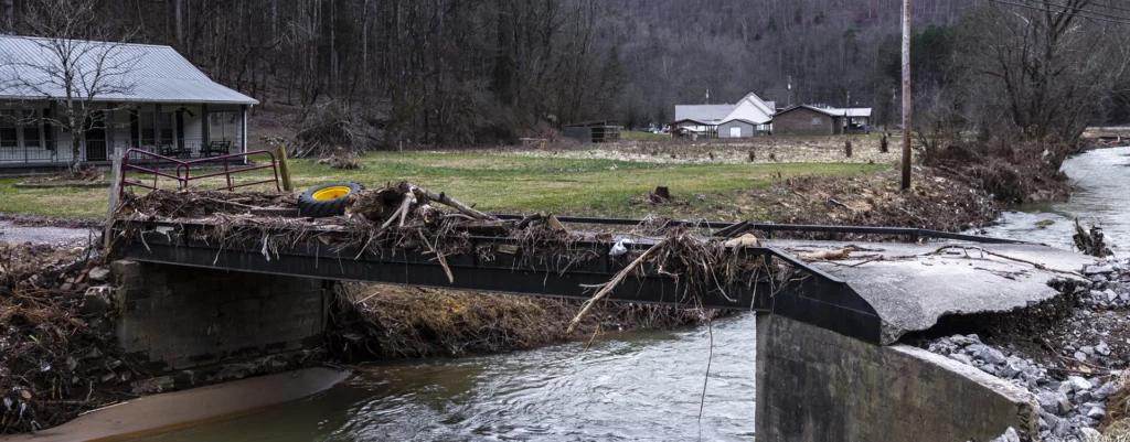 Kentucky flooding damage showing a rural bridge washed out and covered with debris near homes along a creek.