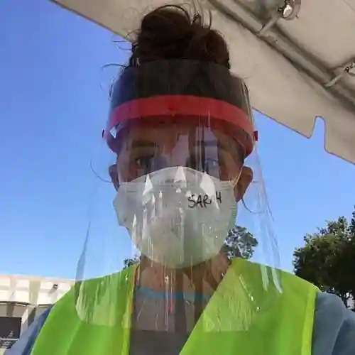 A volunteer wearing a neon safety vest, N95 mask, and clear face shield stands outside under a canopy during COVID-19 response efforts.