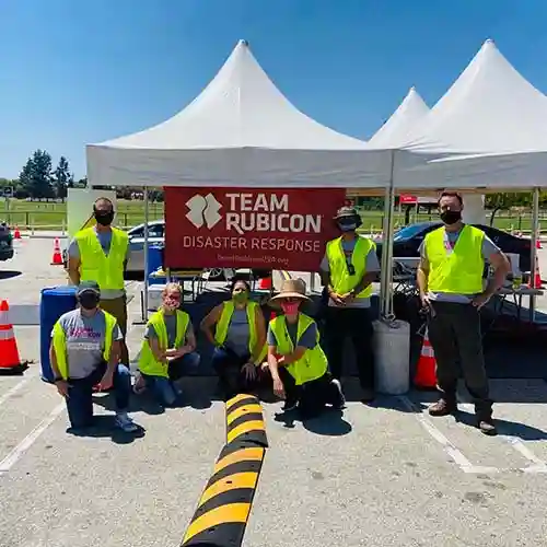 group of masked volunteers in yellow safety vests kneel and stand in front of a Team Rubicon Disaster Response tent in a parking lot.