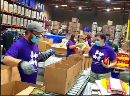 lunteers in purple All Hands and Hearts shirts and face masks pack canned goods and supplies into boxes on a conveyor belt in a warehouse.