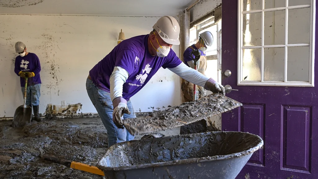 Volunteers in purple All Hands and Hearts shirts and hard hats clean out a mud-filled home, shoveling debris into wheelbarrows after a flood.