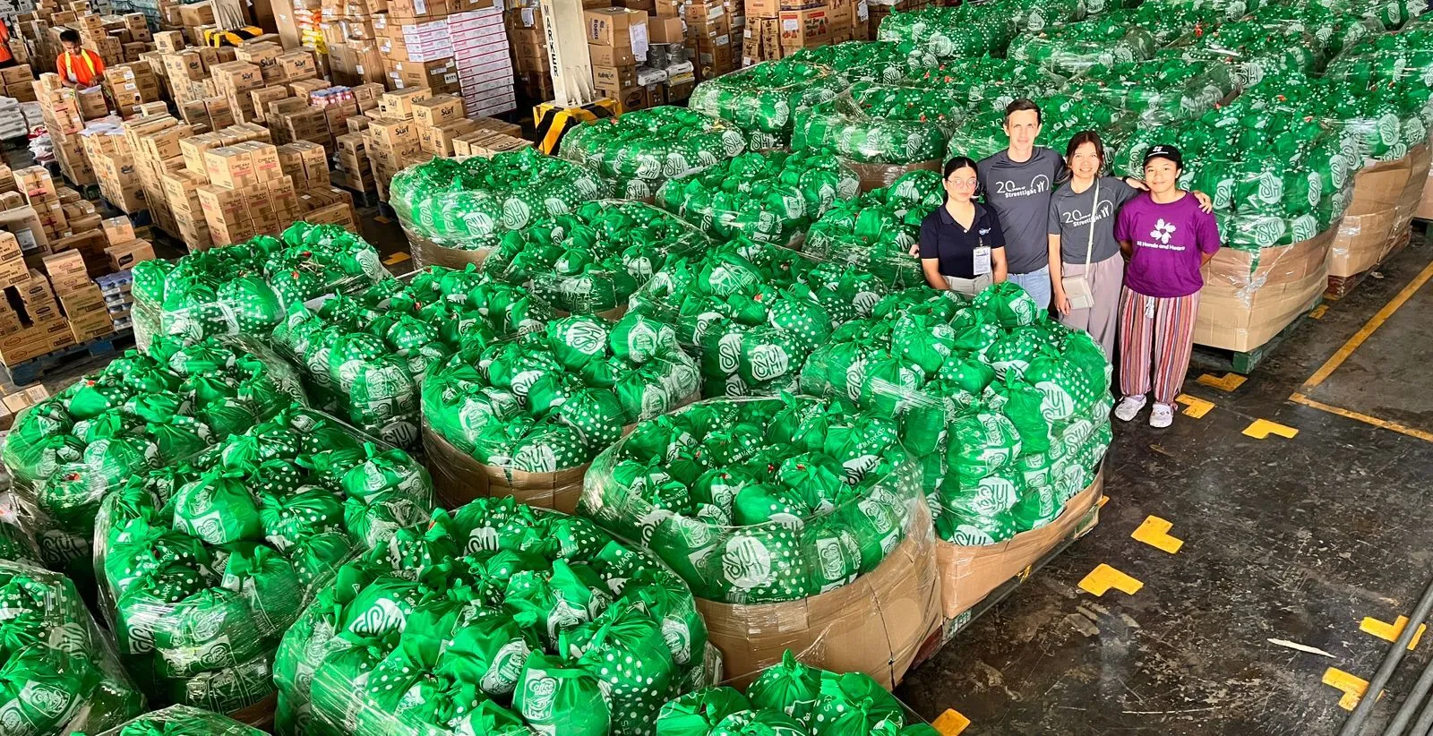 Four individuals stand amongst hundreds of bags containing relief items